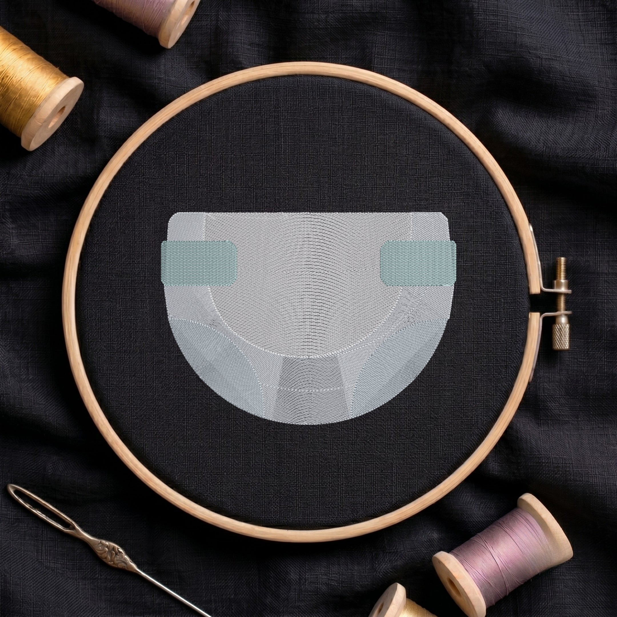 An embroidery design of a diaper in light gray and teal thread, held in a wooden embroidery hoop on a dark fabric background, surrounded by spools of thread and a needle.