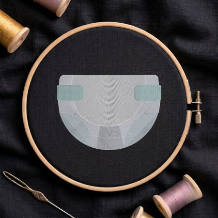 An embroidery design of a diaper in light gray and teal thread, held in a wooden embroidery hoop on a dark fabric background, surrounded by spools of thread and a needle.