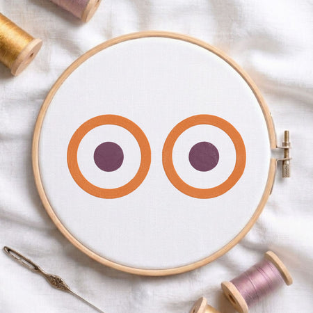 A circular embroidery hoop holding white fabric with two stylized owl-like eye motifs in orange, white, and dark purple. The hoop is styled on a white fabric background with spools of thread and a needle around it.
