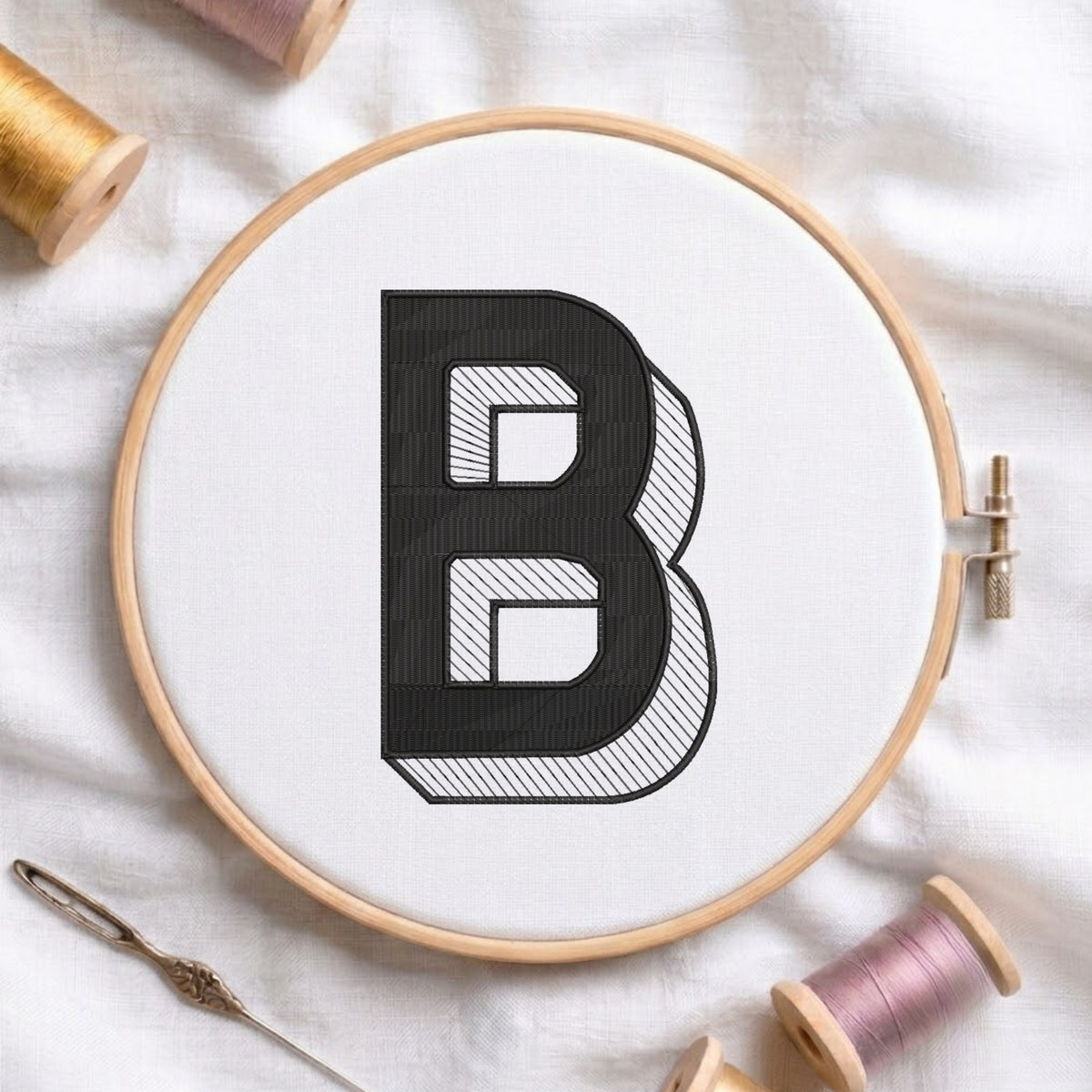 A wooden embroidery hoop holding white fabric with a large black letter B stitched in a bold, patterned style. The hoop is photographed on a white fabric surface with spools of thread and a needle around it.