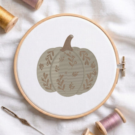 A circular embroidery hoop holding white fabric with a stitched pumpkin design in muted beige and brown tones. The hoop is styled on a white fabric surface with spools of thread and a needle around it.