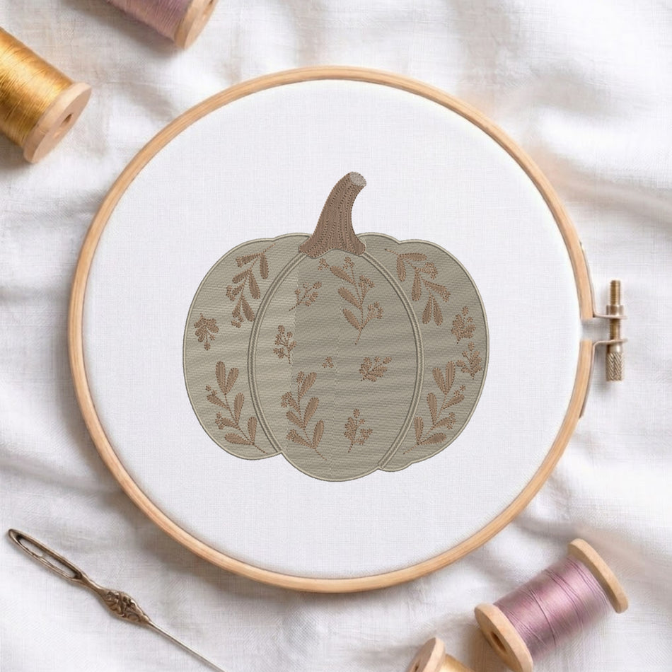 A circular embroidery hoop holding white fabric with a stitched pumpkin design in muted beige and brown tones. The hoop is styled on a white fabric surface with spools of thread and a needle around it.