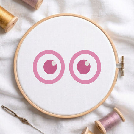 A circular embroidery hoop holding white fabric with a simple cartoon eye design in pink and magenta, surrounded by spools of thread and a needle on a white fabric background.