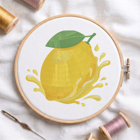 The lemon embroidery design centered in a wooden embroidery hoop, displayed with embroidery thread spools and a needle on a white fabric background.