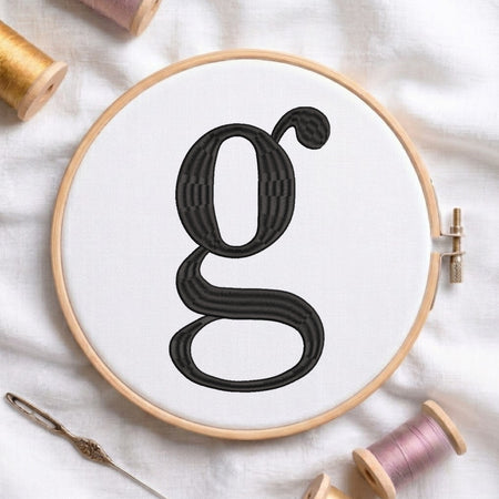 A wooden embroidery hoop holding white fabric with a large black embroidered letter 'g'. Spools of thread and a needle are arranged around it on a white fabric background.