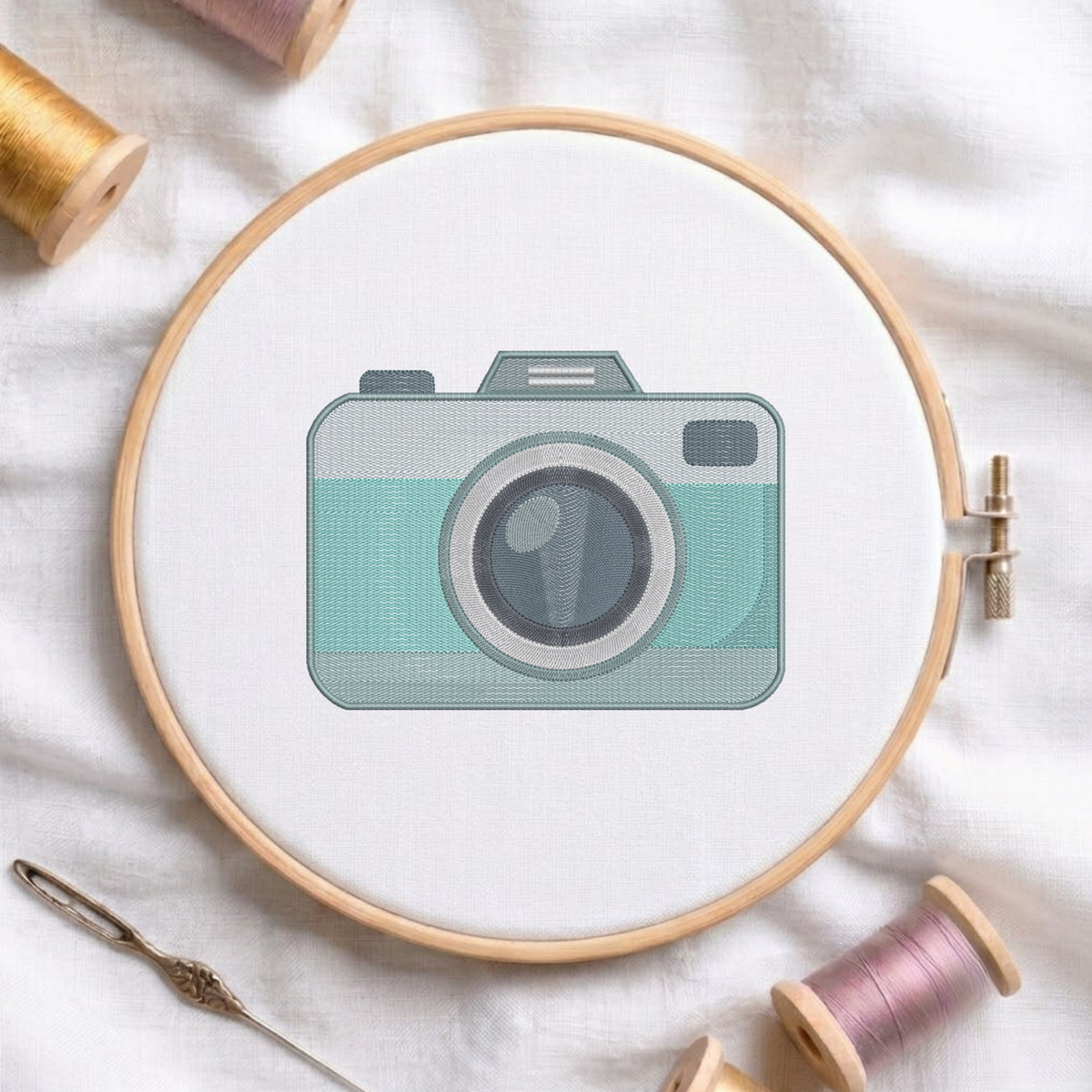 A circular embroidery hoop holding white fabric with a stylized camera design in mint green, gray, and black. The hoop is photographed on white fabric with spools of thread and a needle around it.