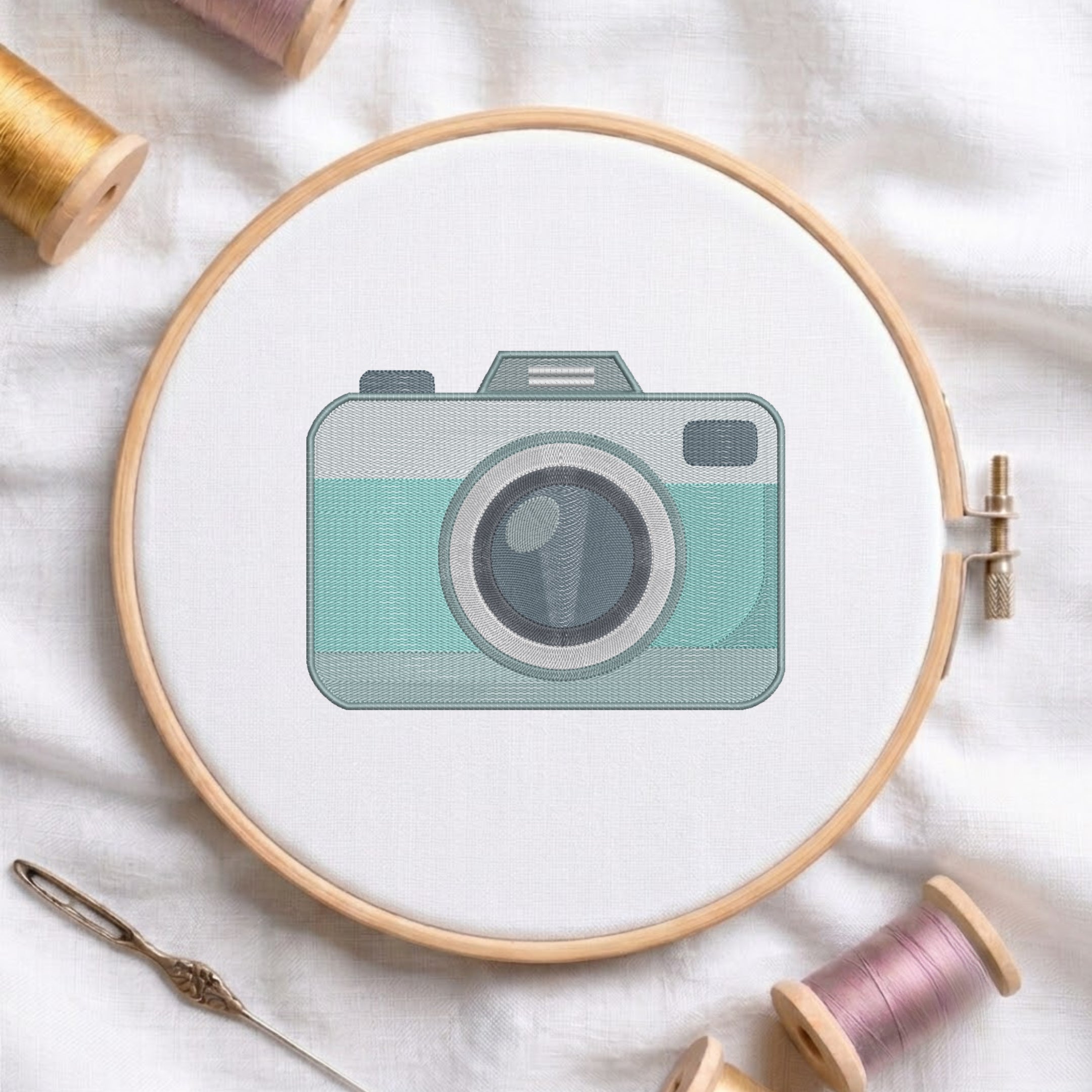 A circular embroidery hoop holding white fabric with a stylized camera design in mint green, gray, and black. The hoop is photographed on white fabric with spools of thread and a needle around it.