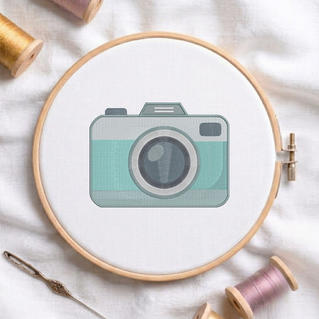 A circular embroidery hoop holding white fabric with a stylized camera design in mint green, gray, and black. The hoop is photographed on white fabric with spools of thread and a needle around it.