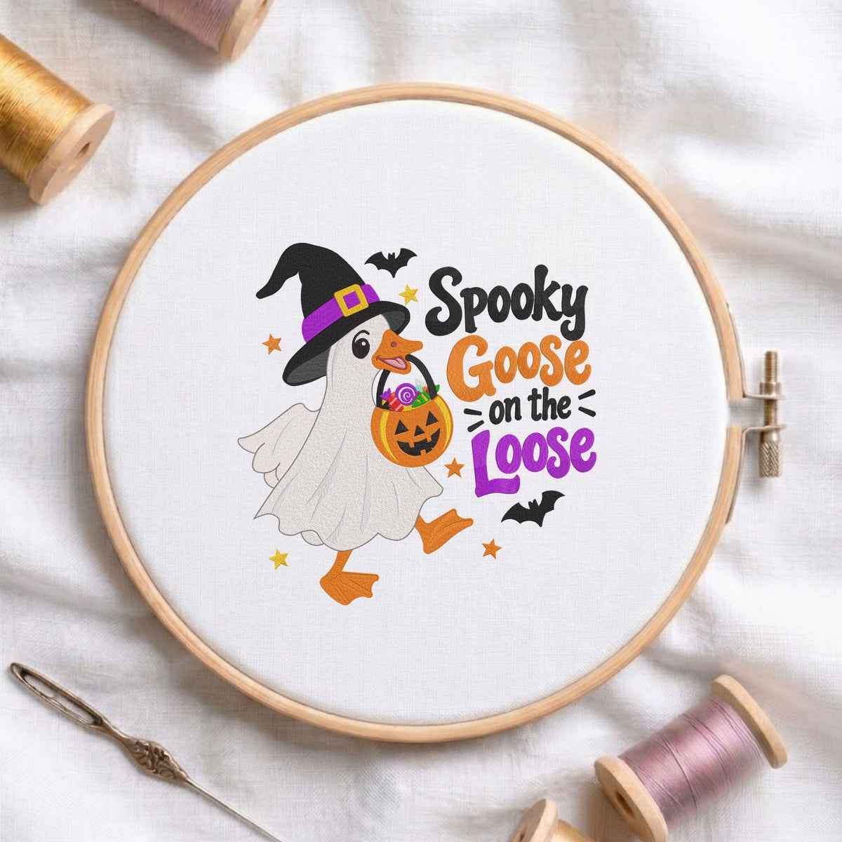 Spooky Goose on the Loose" funny ghost goose wearing a witch hat and holding a jack-o-lantern candy bucket machine embroidery design shown in a wooden embroidery hoop on white fabric with thread spools and a needle