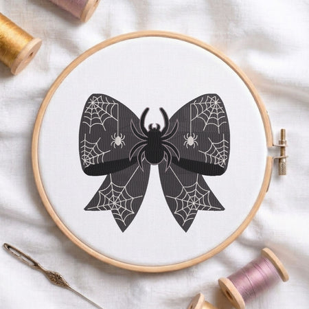 A white fabric stretched in a wooden embroidery hoop, featuring an embroidered design of a dark spiderweb-patterned bow centered with a large black spider.