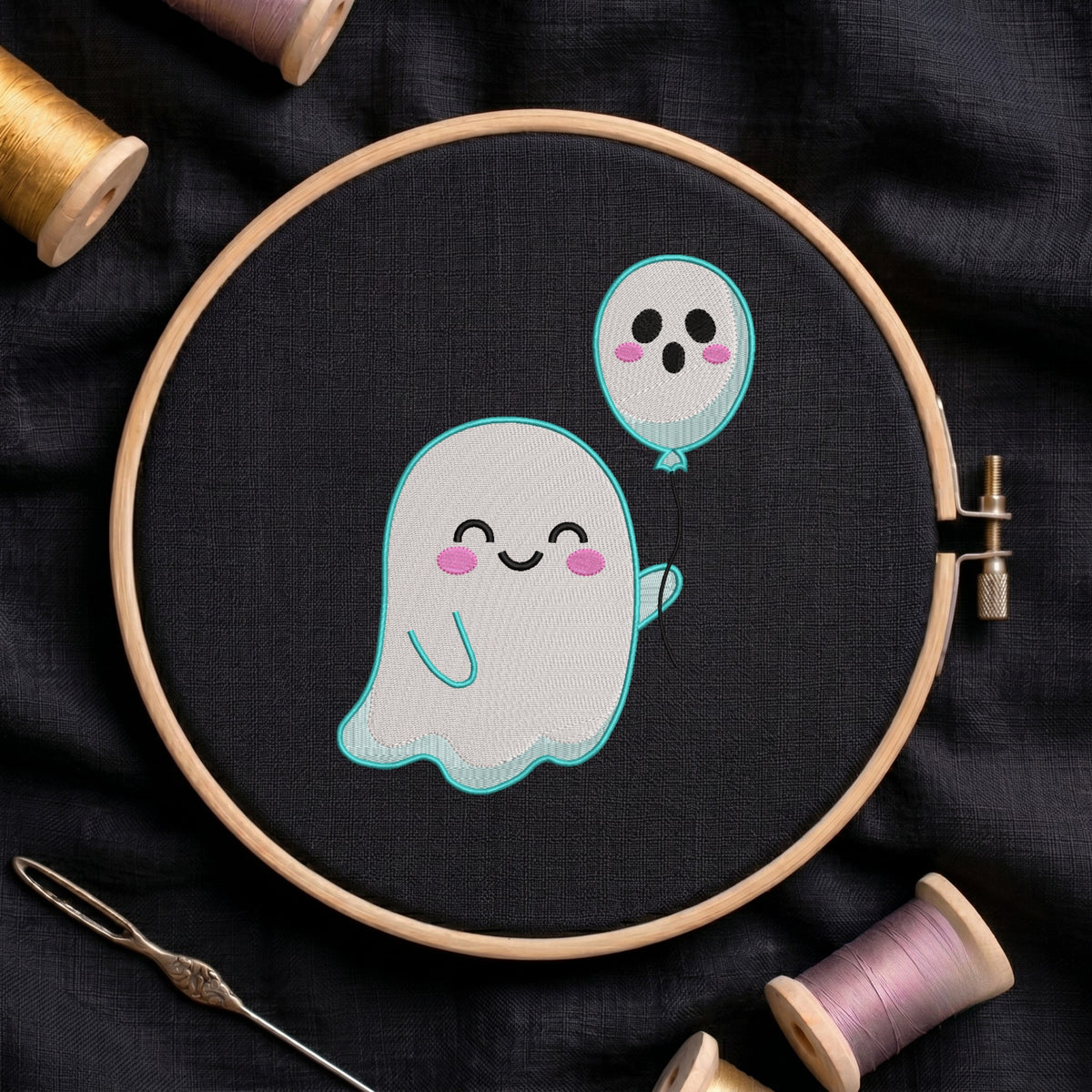 A circular embroidery hoop holding black fabric with a cute embroidered design of a smiling white ghost holding a light blue balloon. The hoop is styled on a dark fabric background with spools of thread and a needle.