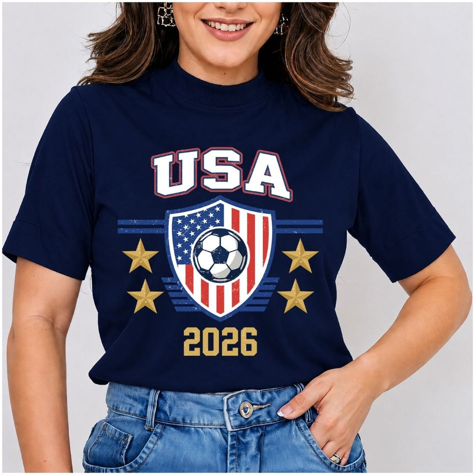 Smiling person wearing a dark blue t-shirt featuring the retro USA 2026 soccer shield graphic and bold typography.