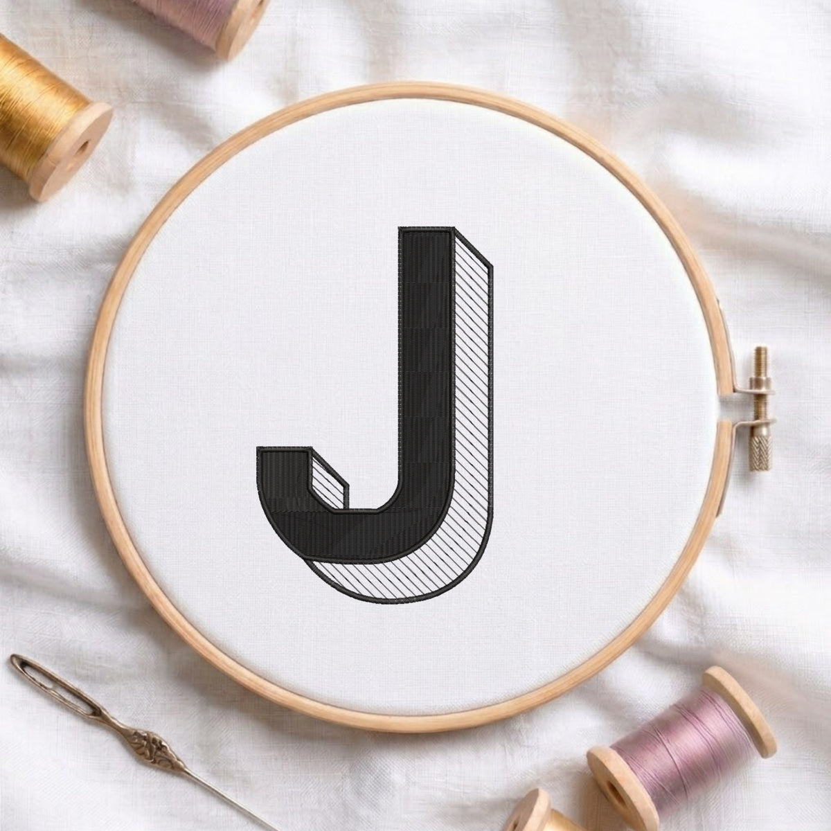 A wooden embroidery hoop holding white fabric with a large black letter J stitched in the center. A needle and thread spools are arranged around it on a white fabric background.