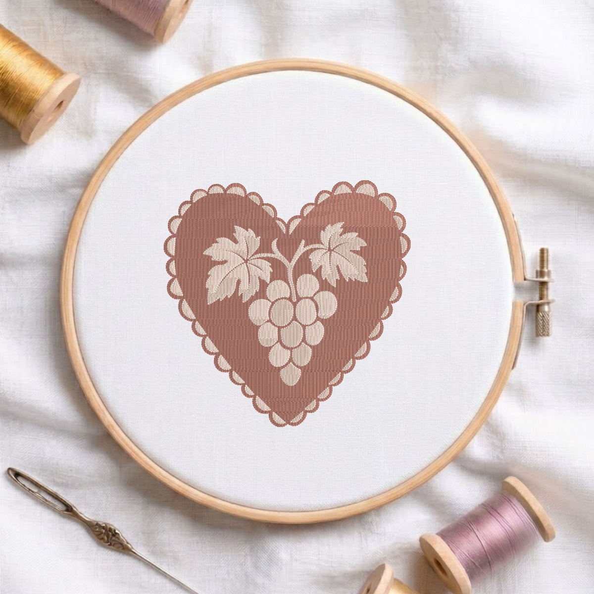 A wooden embroidery hoop holding white fabric with a stitched heart design featuring a brown scalloped border and a cream-colored grape motif. Spools of thread and a needle are arranged around it on a white fabric background.