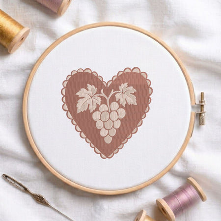 A wooden embroidery hoop holding white fabric with a stitched heart design featuring a brown scalloped border and a cream-colored grape motif. Spools of thread and a needle are arranged around it on a white fabric background.