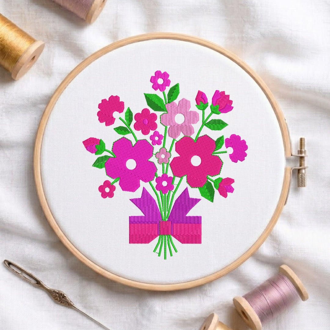 A circular embroidery hoop containing a floral bouquet design in bright pink, magenta, and green on white fabric, styled with spools of thread and a needle on a white cloth background.
