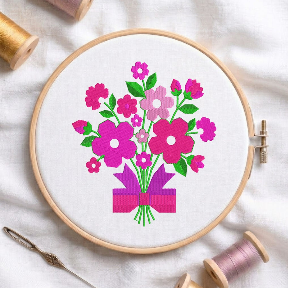 A circular embroidery hoop containing a floral bouquet design in bright pink, magenta, and green on white fabric, styled with spools of thread and a needle on a white cloth background.