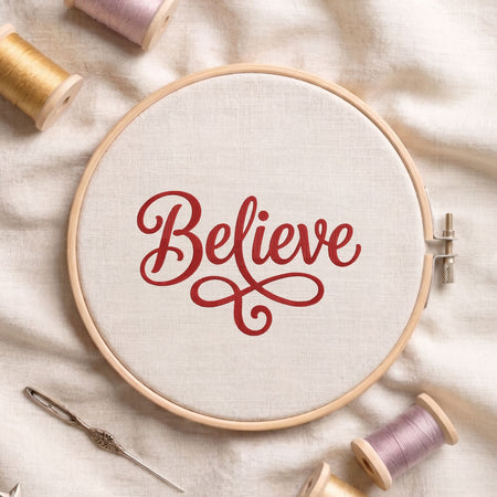 "Believe" word art machine embroidery design with elegant cursive lettering displayed inside a round wooden hoop on white linen fabric, accompanied by wooden thread spools and a needle.