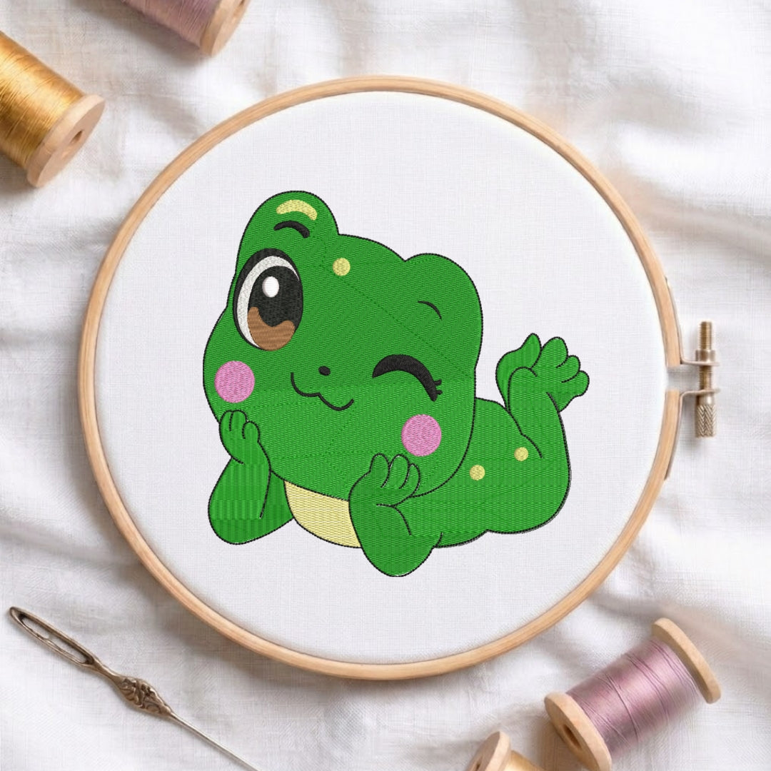 A circular embroidery hoop holding white fabric with a cartoon green frog design, surrounded by spools of thread and a needle on a white fabric background.