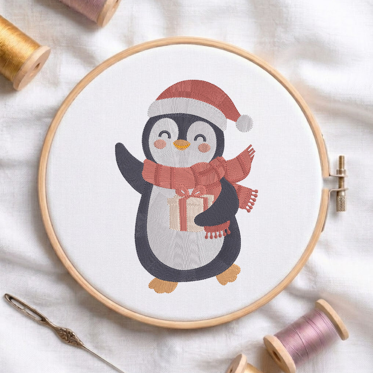 A circular embroidery hoop on white fabric, featuring a stitched penguin wearing a red Santa hat and matching red scarf while holding a gift box. Spools of thread and a needle are arranged around the hoop.