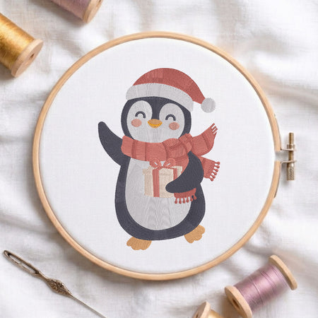 A circular embroidery hoop on white fabric, featuring a stitched penguin wearing a red Santa hat and matching red scarf while holding a gift box. Spools of thread and a needle are arranged around the hoop.