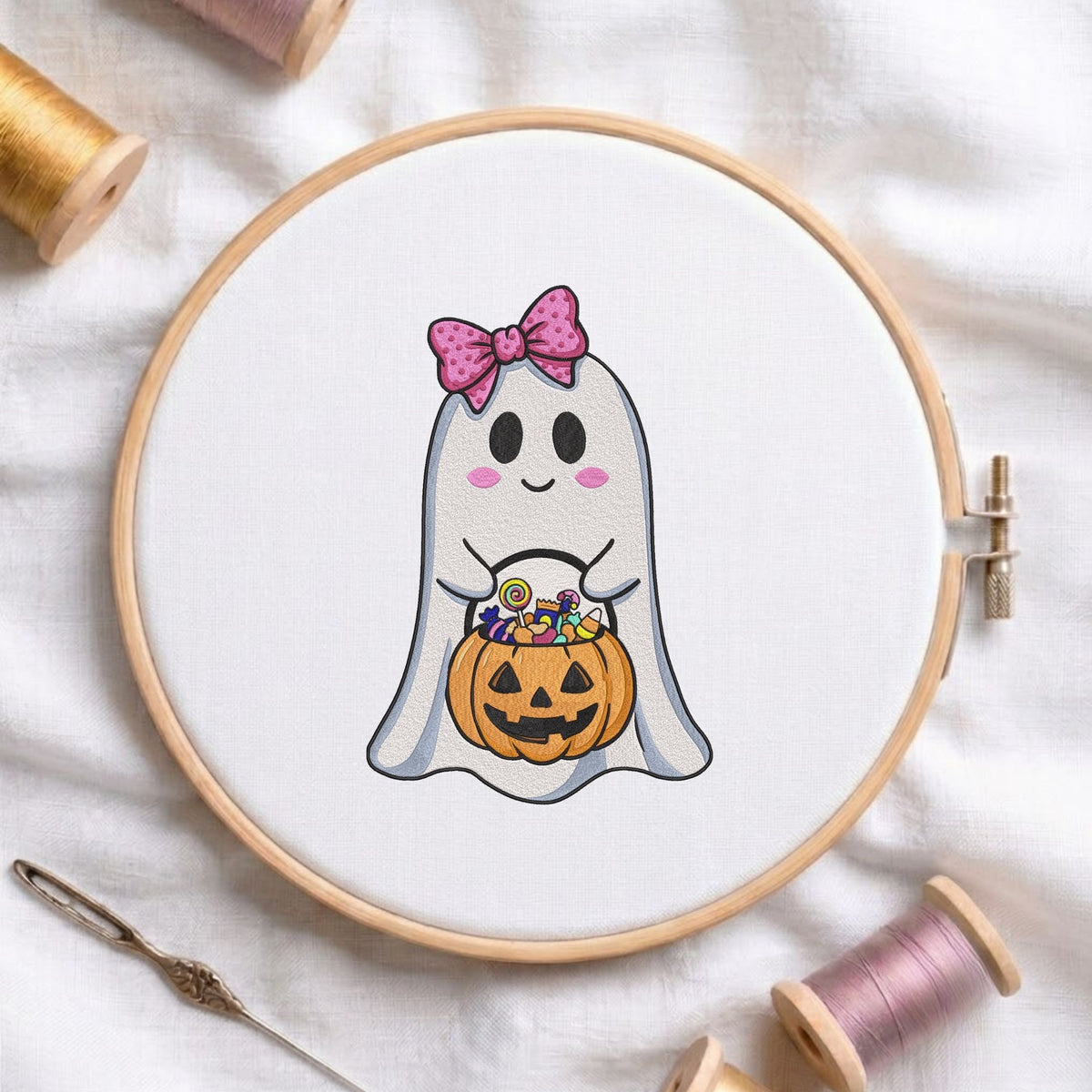Cute ghost holding a candy-filled jack-o'-lantern bucket with a pink polka dot bow machine embroidery design displayed in a wooden embroidery hoop on white fabric with thread spools and a needle