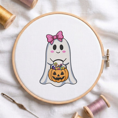 Cute ghost holding a candy-filled jack-o'-lantern bucket with a pink polka dot bow machine embroidery design displayed in a wooden embroidery hoop on white fabric with thread spools and a needle