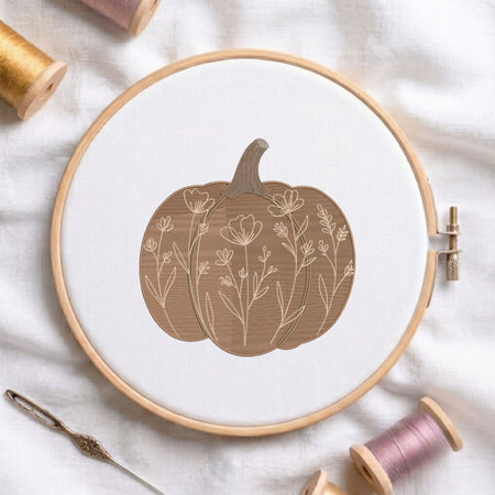 An embroidered beige pumpkin featuring intricate botanical wildflower and stem patterns, stitched inside a wooden hoop on white fabric with sewing threads.