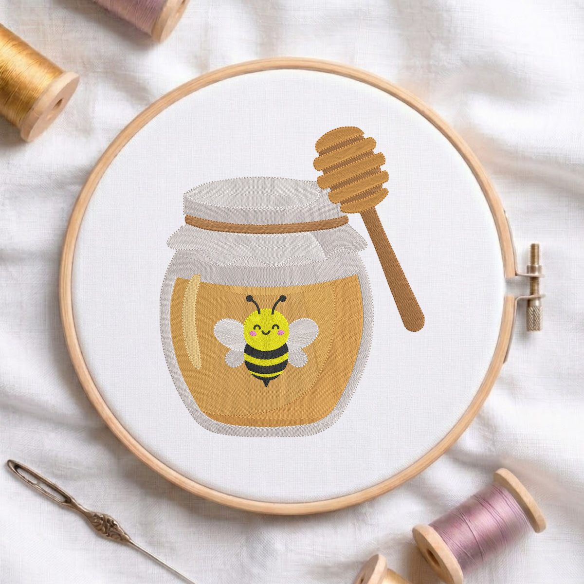 A circular embroidery hoop displays a stitched design of a honey jar with a smiling bee and a honey dipper. The hoop is styled on a white fabric background with spools of thread and a needle.