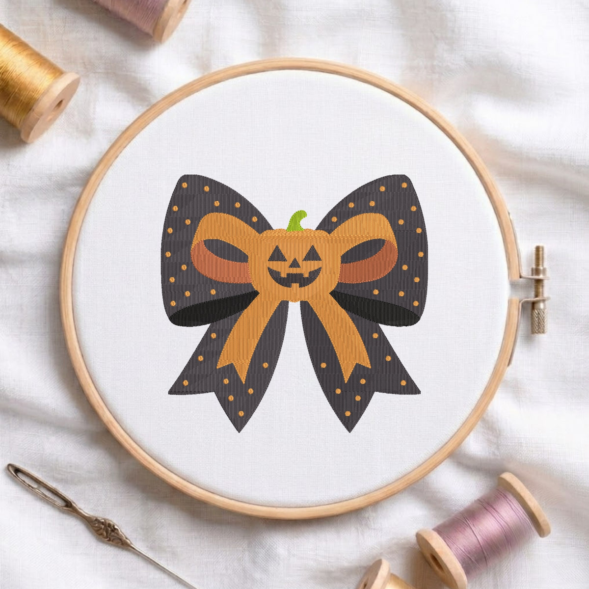 A white fabric stretched in a wooden embroidery hoop, featuring an embroidered design of a dark polka-dot bow centered with a cute jack-o'-lantern pumpkin.