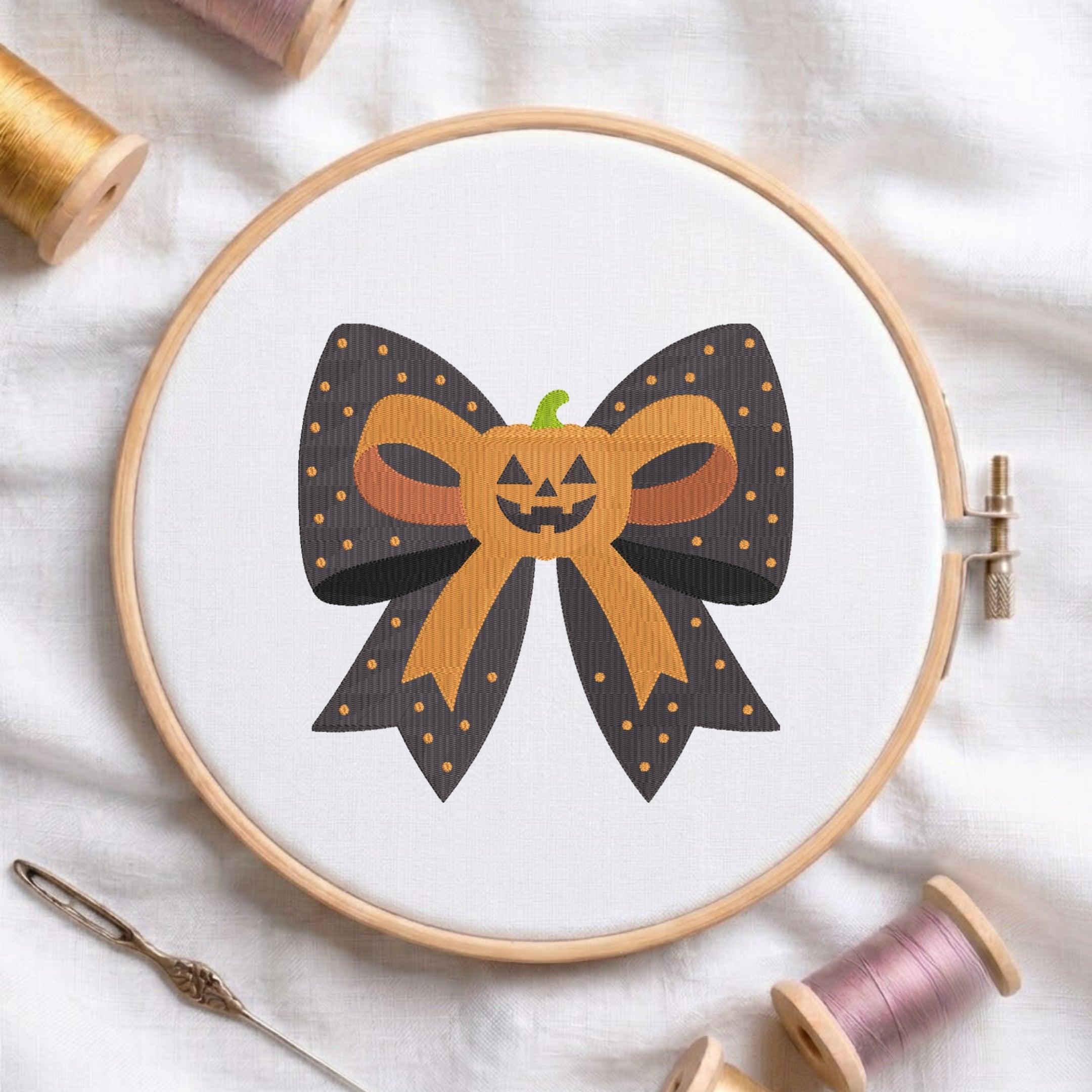 A white fabric stretched in a wooden embroidery hoop, featuring an embroidered design of a dark polka-dot bow centered with a cute jack-o'-lantern pumpkin.