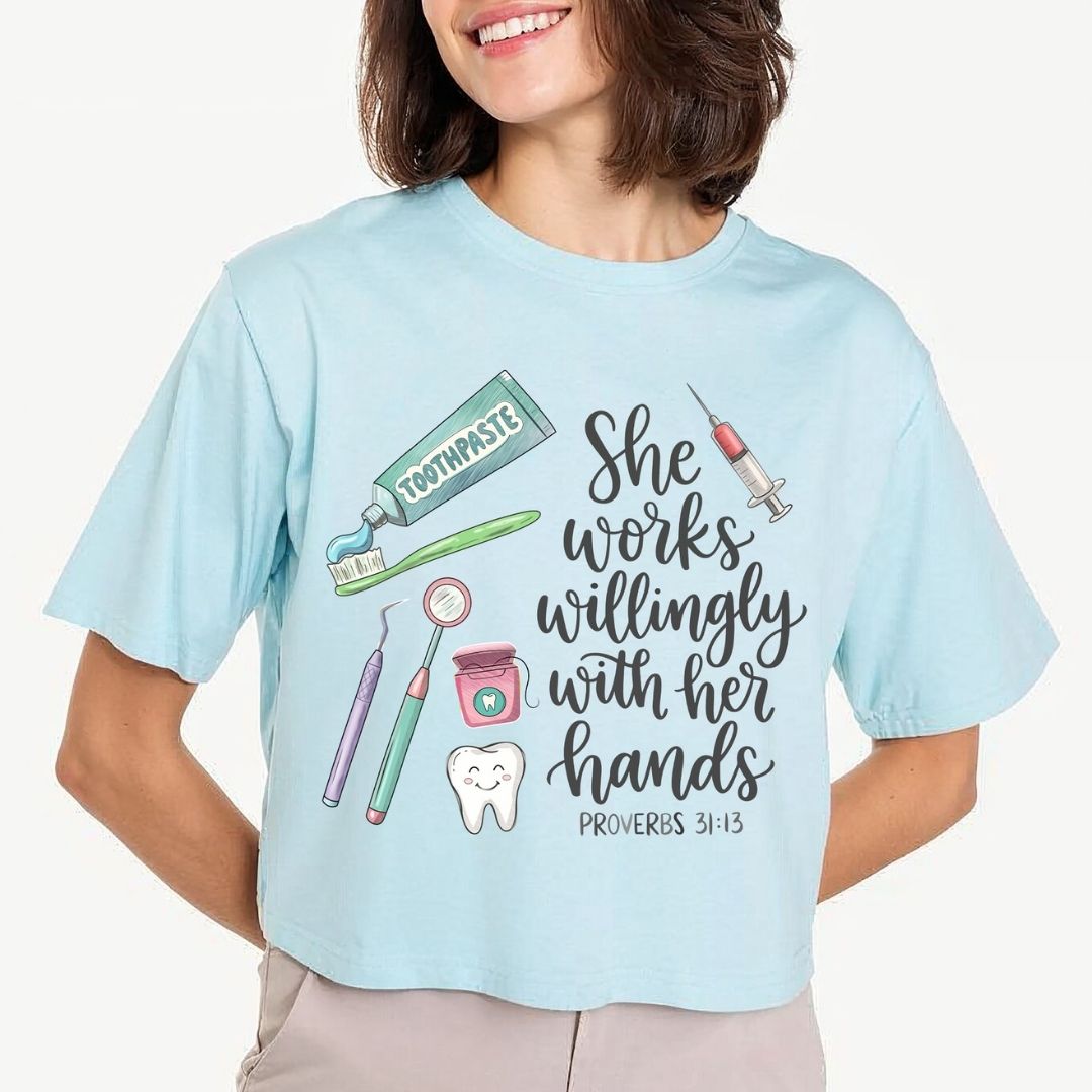 A woman wearing a light blue graphic t-shirt with a dental-themed illustration and the text 'She works willingly with her hands PROVERBS 31:13'.