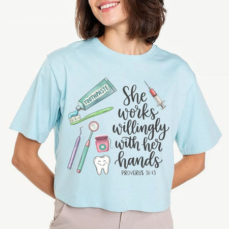 A woman wearing a light blue graphic t-shirt with a dental-themed illustration and the text 'She works willingly with her hands PROVERBS 31:13'.