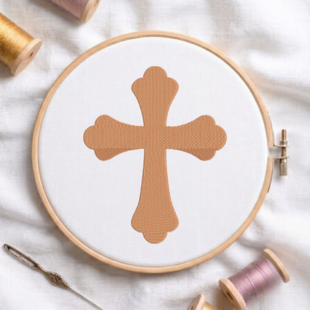 A circular embroidery hoop holding white fabric with a tan cross design, surrounded by spools of thread and a needle on a white fabric background.