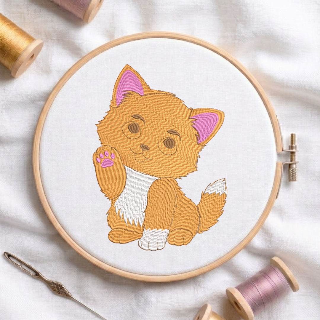 The orange kitten embroidery design secured in a wooden embroidery hoop, displayed with embroidery thread spools and a needle on a white fabric background.