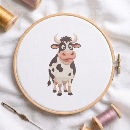 Funny cute cow machine embroidery design shown in a wooden embroidery hoop on white fabric with thread spools and a needle