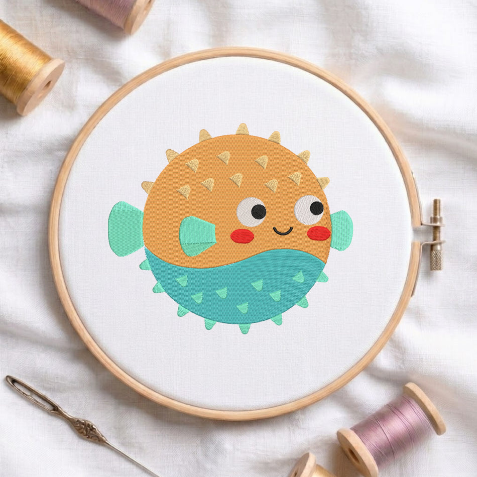 A round embroidery hoop holding white fabric with a colorful cartoon puffer fish design. The fish has an orange top half with small yellow spikes, a teal bottom half with green triangles, a teal fin, and red cheeks.