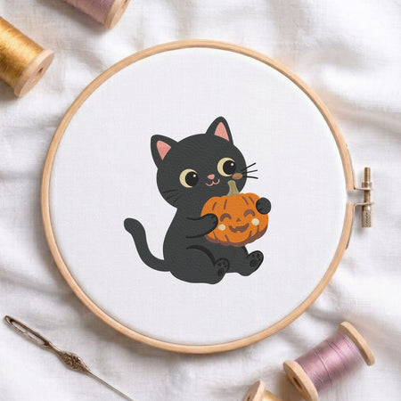 Cute black cat hugging a mini jack-o-lantern pumpkin Halloween machine embroidery design shown in a wooden embroidery hoop on white fabric with thread spools and a needle