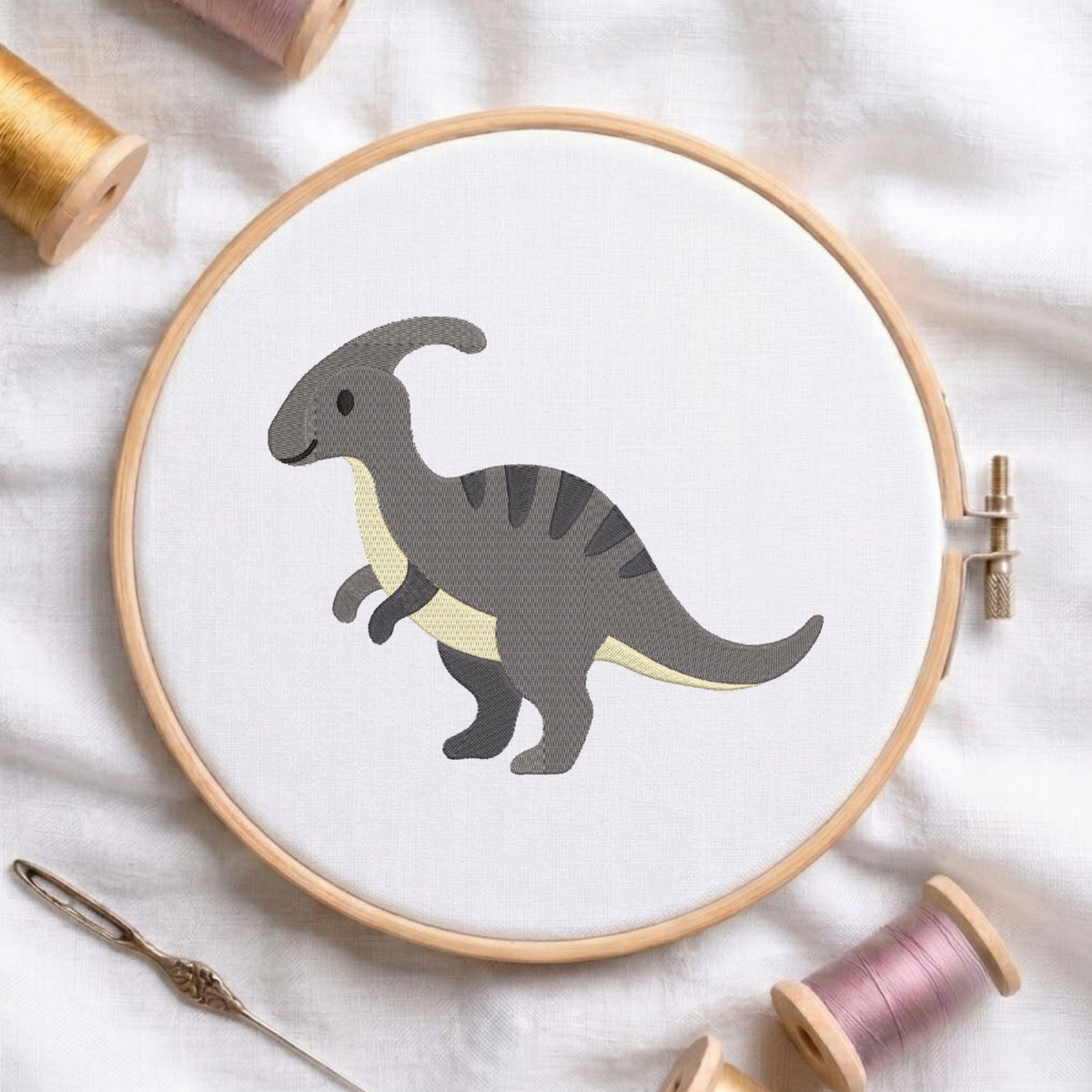 A circular embroidery hoop holding white fabric with a stylized gray dinosaur design, surrounded by spools of thread and a needle on a white fabric background.