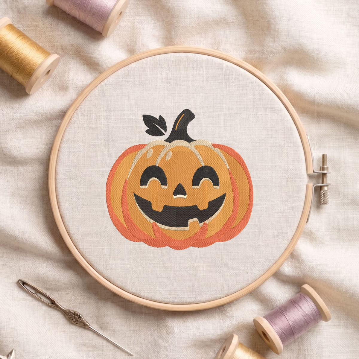 Jack-o'-lantern pumpkin machine embroidery design featuring a smiling carved face and green leaf stem, displayed inside a round wooden hoop on white linen fabric alongside thread spools and a needle.