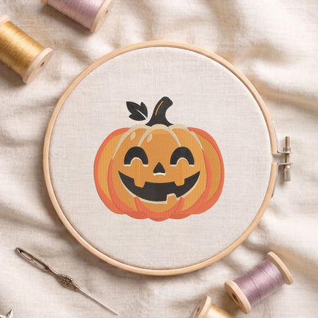 Jack-o'-lantern pumpkin machine embroidery design featuring a smiling carved face and green leaf stem, displayed inside a round wooden hoop on white linen fabric alongside thread spools and a needle.