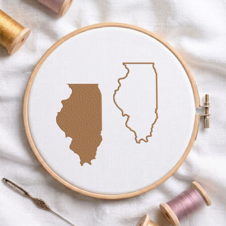 A wooden embroidery hoop on white fabric, showing a tan Illinois state silhouette filled with texture and a matching outline. Thread spools, a needle, and a small tool are arranged around the hoop.