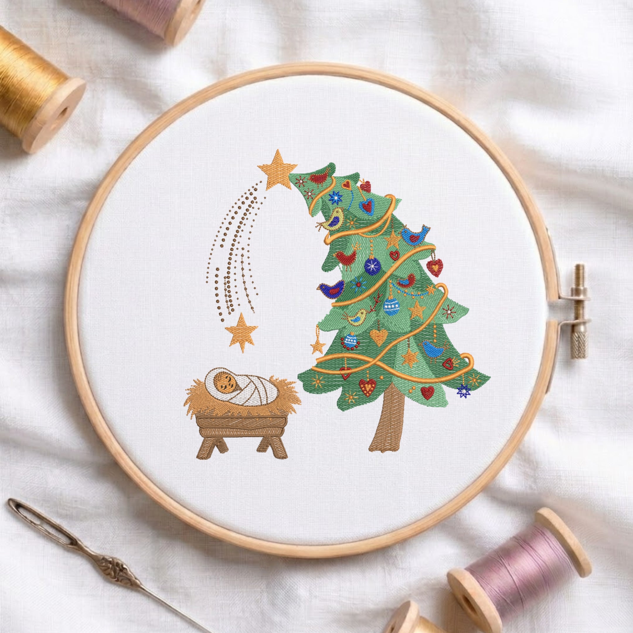 A circular embroidery hoop on white fabric displays a Christmas nativity design: a green decorated tree, gold stars, birds, and a baby wrapped in white cloth in a wooden manger. Spools of thread, a needle, and soft fabric surround the hoop.