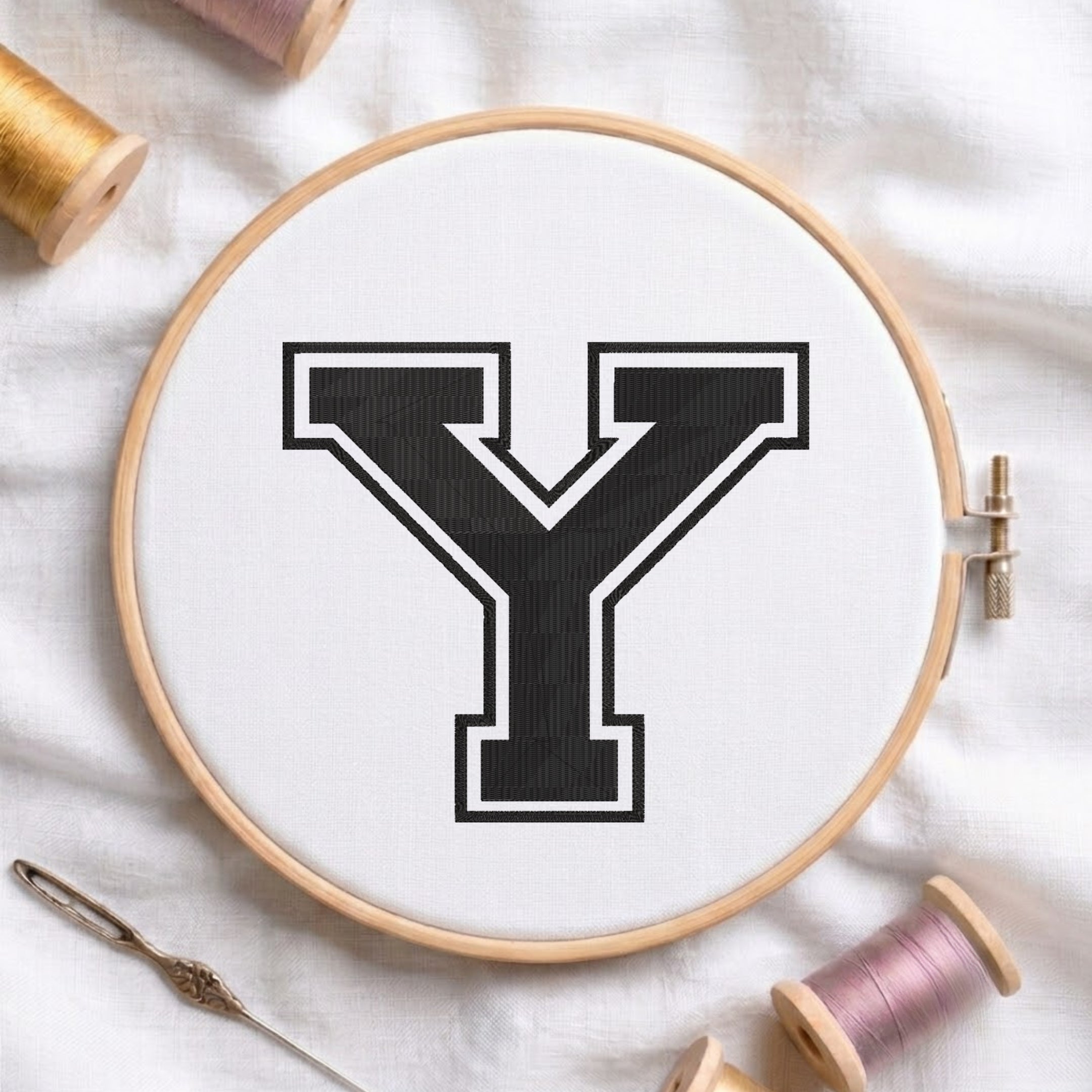 A wooden embroidery hoop holding white fabric with a large black letter Y outlined in white, surrounded by spools of thread and a needle on a white fabric background.