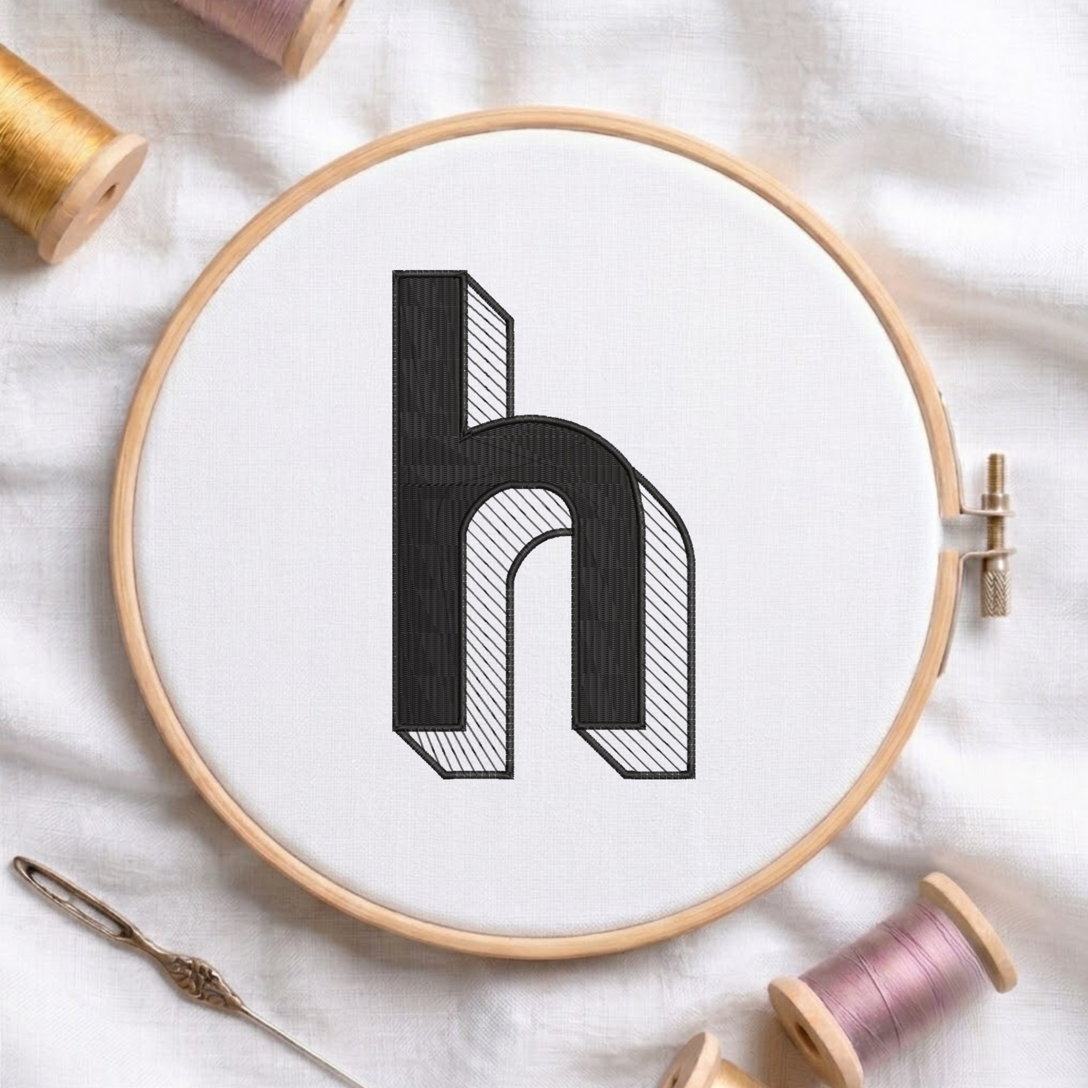 A wooden embroidery hoop holds white fabric with a large black embroidered letter 'h' shown in a 3D block style. The hoop is styled on a white fabric background with spools of thread and a needle.