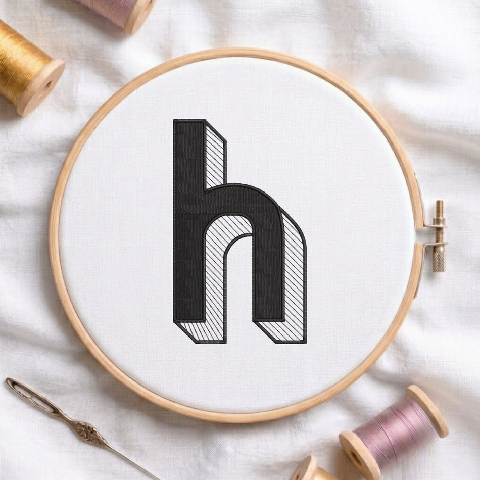 A wooden embroidery hoop holds white fabric with a large black embroidered letter 'h' shown in a 3D block style. The hoop is styled on a white fabric background with spools of thread and a needle.