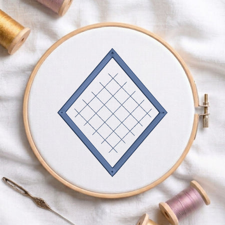 A wooden embroidery hoop holding white fabric with a blue diamond grid design, styled on a white cloth with spools of thread and a needle around it.