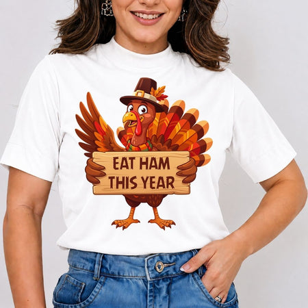 A woman wearing a white short-sleeve t-shirt with a cartoon turkey graphic holding a wooden sign that reads 'EAT HAM THIS YEAR'. The woman is also wearing blue jeans, and the background is a plain light gray studio wall.