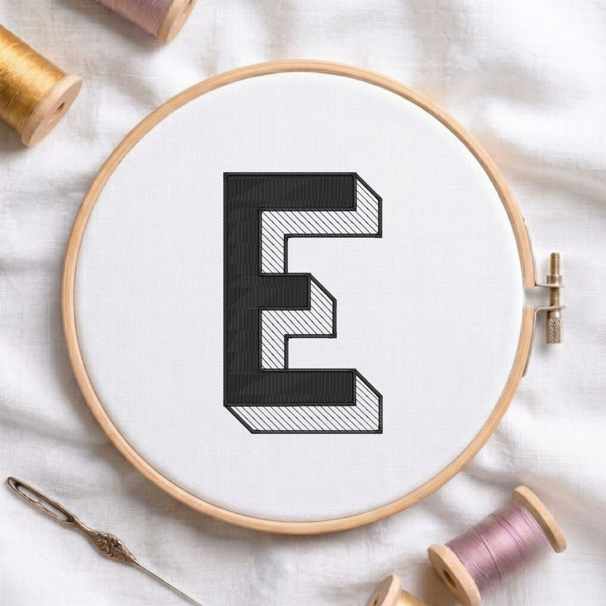 A wooden embroidery hoop holding white fabric with a large black letter E design, surrounded by spools of thread and a needle on a white fabric background.