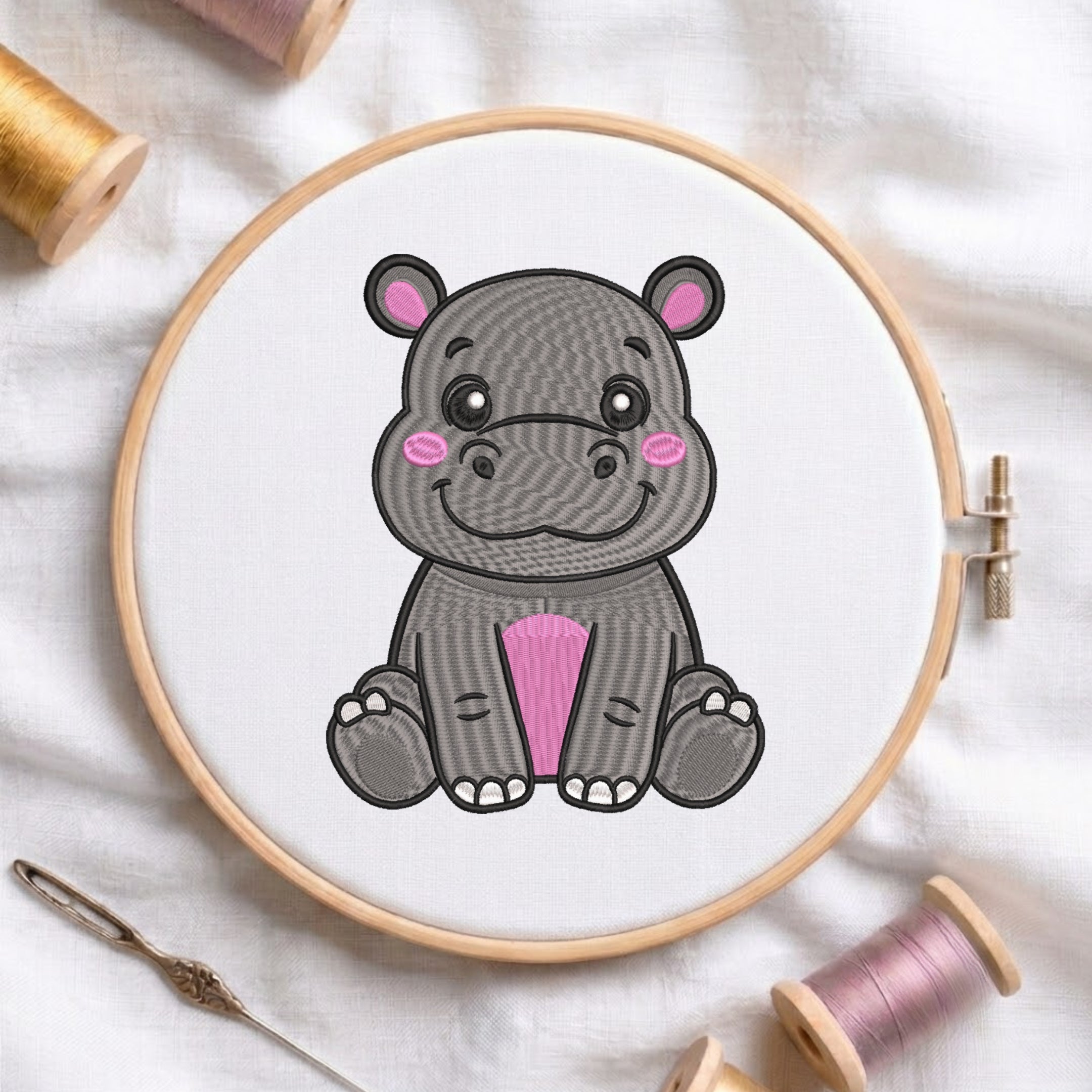 A wooden embroidery hoop holding white fabric with a cartoon hippo design in gray, black, pink, and white. The hoop is photographed on a white fabric surface with spools of thread and a needle around it.
